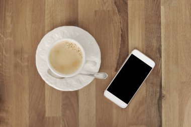 Boş siyah ekran lı beyaz cep telefonunun görüntüsünü ve ahşap masa üzerinde bir fincan kahve, üstteki görünümü ile alay