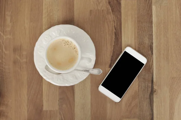 Изображение белого мобильного телефона с пустым черным экраном, чашка кофе на деревянном столе, вид сверху — стоковое фото