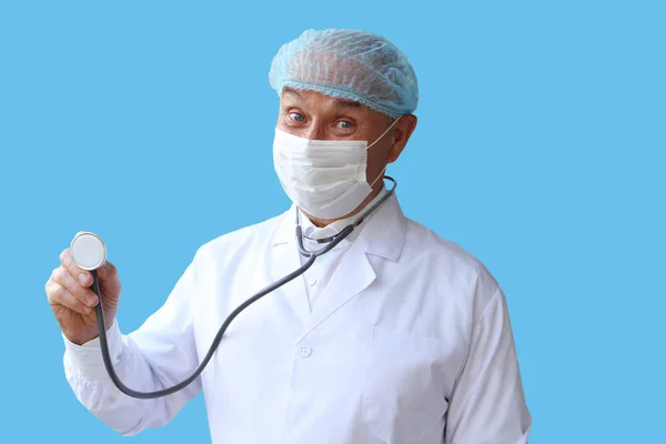 Medico maschio in un cappotto bianco, cappuccio e maschera sta tenendo uno stetoscopio nella mano destra su uno sfondo blu, isolare — Foto Stock