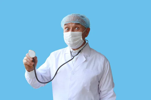 Medico maschio in un cappotto bianco, cappuccio e maschera tiene uno stetoscopio nella mano destra, occhi buffi goggling, su uno sfondo blu, isolare, copiare spazio — Foto Stock