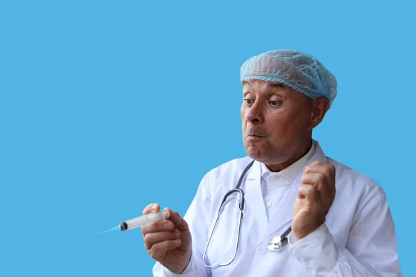 Мужчина врач в белом халате и кепке держит в руке медицинский шприц, имитирующий удивление, на синем фоне, изолировать, копировать пространство — стоковое фото
