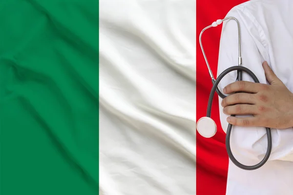 Doctor masculino de bata blanca sosteniendo en su mano un instrumento médico con un estetoscopio en el fondo de la bandera nacional Italia, primer plano, concepto, espacio de copia — Foto de Stock