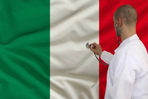 Medico maschio in mantello bianco applicato uno strumento medico con stetoscopio alla bandiera nazionale Italia, vista posteriore, concetto, spazio per la copia — Foto Stock