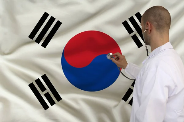 Doctor masculino en bata blanca aplicado un instrumento médico con un estetoscopio a la bandera nacional, vista posterior, concepto, espacio de copia — Foto de Stock