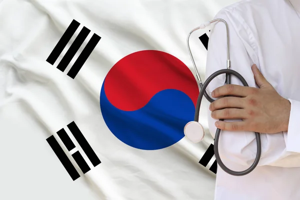 Male doctor in white coat holding in his hand a medical instrument with a stethoscope against the background of the national flag, close-up, concept, copy space — Stock Photo, Image