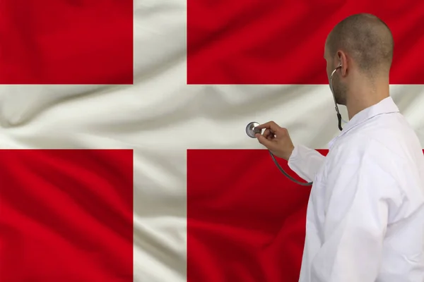 Male doctor in white coat applied a medical instrument with a stethoscope to the national flag, back view, concept, copy space — Stock Photo, Image