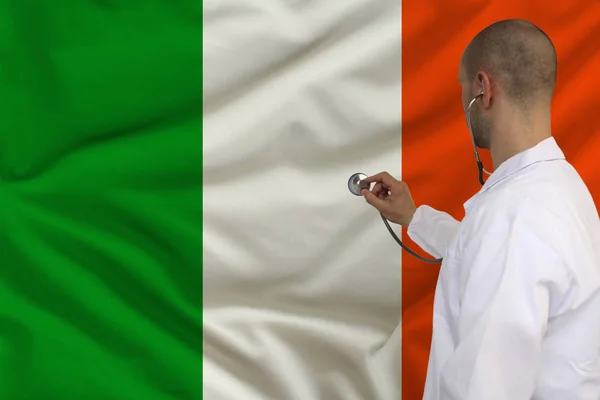 Doctor masculino en bata blanca aplicado un instrumento médico con un estetoscopio a la bandera nacional, vista posterior, concepto, espacio de copia — Foto de Stock
