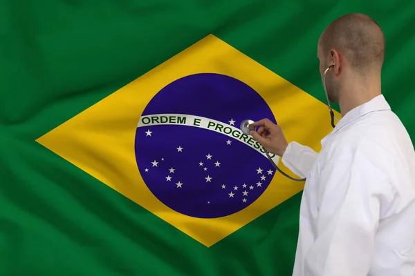 Medico maschio in camice bianco applicato uno strumento medico con uno stetoscopio alla bandiera nazionale brasiliana, vista posteriore, concetto, spazio copia — Foto Stock