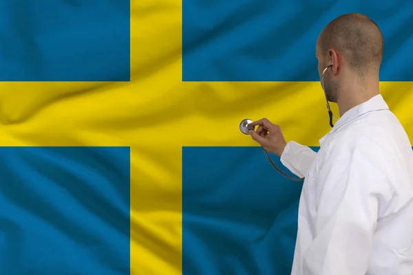 Mannelijke arts in witte vacht toegepast een medisch instrument met een stethoscoop aan de nationale vlag Zweden, achteraanzicht, concept, kopie ruimte — Stockfoto