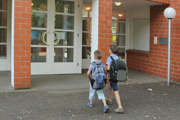 Dois jovens estudantes com mochilas se aproximam da entrada para a escola de tijolo vermelho, o conceito é novamente uma escola — Fotografia de Stock