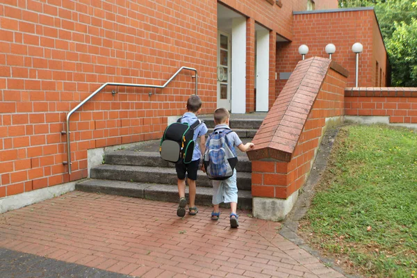 Два младших школьника с рюкзаками ходят в школу, встают, держатся за руки, на крыльце здания школы красного кирпича, концептуальная школа — стоковое фото