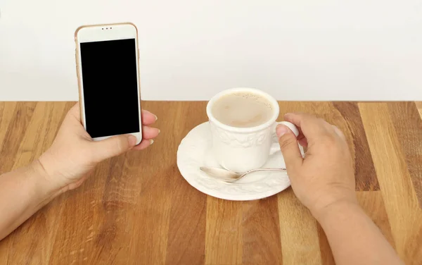 mock up image of female hands holding a white mobile phone with a blank black screen and a cup of coffee