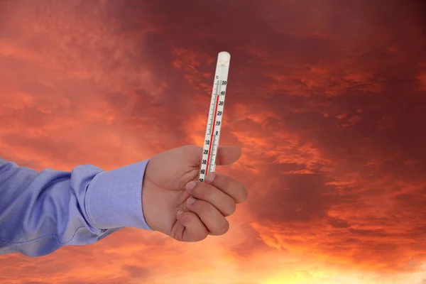 Die Hand des Mannes im Hemd hält ein Thermometer, das die Temperatur misst, das Konzept der Hitze, des Klimawandels, der globalen Erwärmung, 40 Grad Celsius, 120 Grad Fahrenheit gegen den Feuerhimmel bei Sonnenuntergang. — Stockfoto