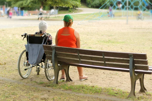 Женщина в оранжевой куртке и пожилая женщина инвалиды в инвалидном кресле, сидеть вместе в парке летом — стоковое фото