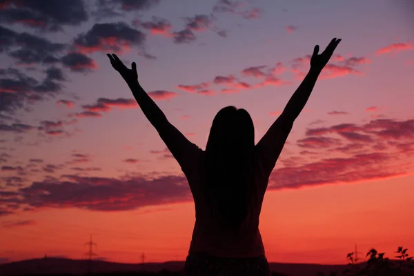 Dark silhouette of a woman in a long dress that raised both hands up to the sky with a beautiful pink sunset with purple clouds, close-up, concept — Stock Photo, Image