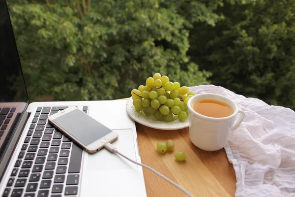노트북과 스마트폰이 테라스의 나무 탁자 위에 놓여 있고, 근처의 차와 포도가 있는 흰색 컵, 정원 밖의 컴퓨터의 프리랜서 작업장 — 스톡 사진