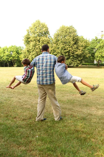 Hombre vestido de verano se convierte en sus dos hijos, jugando, en el parque, el concepto de una familia feliz, vista trasera. — Foto de Stock