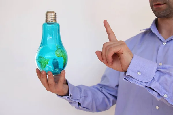 Homem de negócios em uma camisa azul segura em sua mão uma grande lâmpada de vidro azul com um modelo do planeta dentro, mostra um dedo, o conceito de eletricidade, a ideia, ecologia, espaço de cópia — Fotografia de Stock