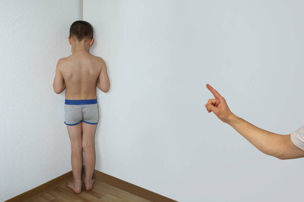 punished boy, a junior schoolboy, standing in a corner undressed, facing the wall, the hand of a parent faces a finger for misconduct, parenting concept