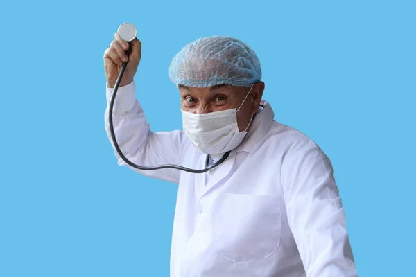 Médico masculino em um casaco branco, tampa e máscara mantém um estetoscópio em sua mão direita, fica em uma pose ameaçadora, em um fundo azul, isolar, espaço de cópia — Fotografia de Stock