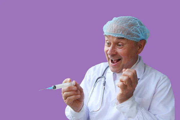 Мужчина врач в белом халате и кепке держит в руке медицинский резиновый шприц, безрассудно улыбается, на фиолетовом фоне, изолировать — стоковое фото