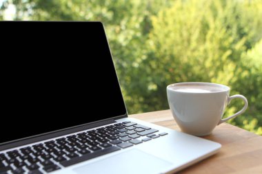 Doğada ahşap bir masada bir fincan kahve ile boş siyah masaüstü ekran ile dizüstü bilgisayar kullanarak, uzak çalışma kavramı mock up