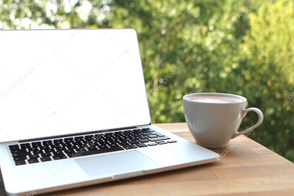 Mock up image of a pleasant morning using a laptop with a blank white desktop screen with a cup of coffee on a wooden table in an outdoor cafe