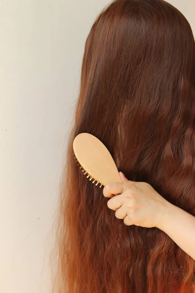 Дівчина з довгим темно-червоним волоссям розчісує їх дерев'яною гребінкою, вертикальною, крупним планом — стокове фото