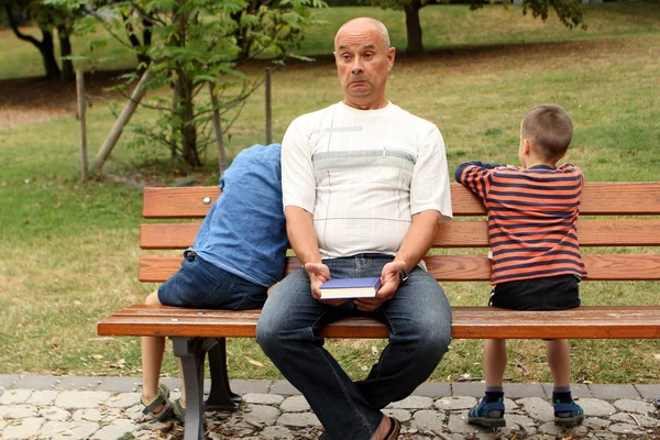 Niños, preescolar, estudiante de primaria están sentados en un banco en el parque al lado del abuelo con un libro, no quieren leer, rechazado, el concepto de relaciones familiares intergeneracionales —  Fotos de Stock