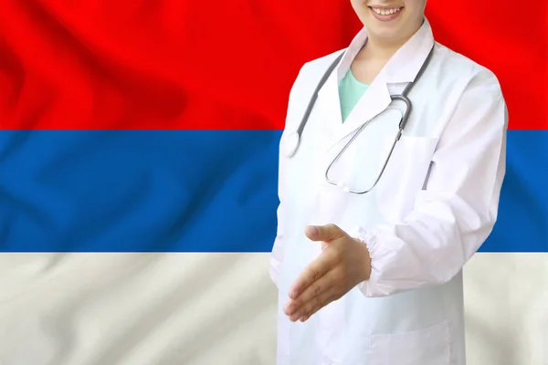 Joven hermosa niña médico con una sonrisa abierta extiende su mano para saludar en el fondo de la bandera nacional, conceptos de atención médica, medicina en serbia — Foto de Stock