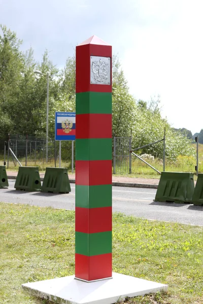 Прикордонний смугастий стовп з державним емблемою на кордоні між Росією і Польщею, на задньому плані прапора з написом Російською Федерацією — стокове фото