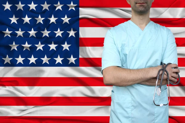 Mannelijke arts in uniform professionele kleding met een stethoscoop staat tegen de achtergrond van de nationale vlag, het concept van de gezondheidszorg en ziektekostenverzekering van het land, close-up, kopieerruimte — Stockfoto