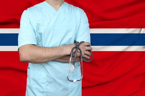 Mannelijke arts in uniform professionele kleding met een stethoscoop staat tegen de achtergrond van de nationale vlag, het concept van de gezondheidszorg en ziektekostenverzekering van het land, close-up, kopieerruimte — Stockfoto