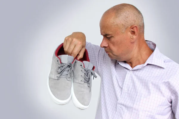 Man in a light shirt holds a pair of gray male moccasins in his hand and sniffs it, a grimace of disgust from the bad smell on his face, close-up, copy space — Stock Photo, Image