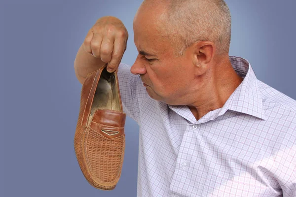 Человек в светлой рубашке держит коричневую мужскую обувь в руке и нюхает ее, гримасу отвращения от плохого запаха на его лице, крупным планом, скопировать пространство — стоковое фото
