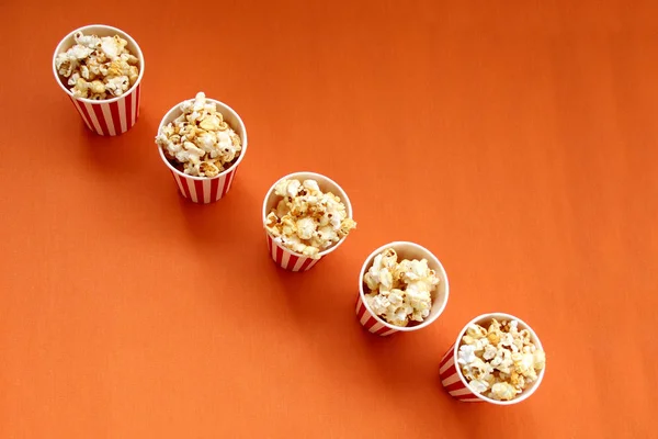 Cinco tazas de papel rojo a rayas, blanco con palomitas de maíz en una fila sobre un fondo naranja brillante, primer plano, espacio de copia, concepto de comida rápida — Foto de Stock