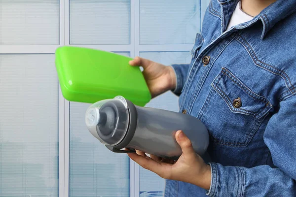 Colegial en una chaqueta de mezclilla sostiene una lonchera de plástico y una botella de agua en sus manos —  Fotos de Stock