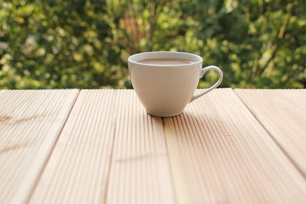 White cup with coffee on a light wooden table against a green summer garden, close-up, copy space — Stock Photo, Image