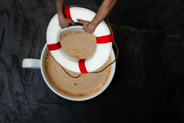 Taza blanca con capuchino y dulces sobre un fondo negro de piedra, manos sosteniendo una boya salvavidas, vista superior, primer plano, espacio para copiar — Foto de Stock