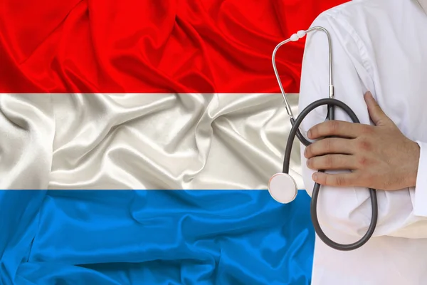 Fotografia parcial do médico em uniforme contra o pano de fundo da bandeira nacional do Luxemburgo em seda brilhante delicada, o conceito de cuidados médicos e seguro no país, close-up, cópia spac — Fotografia de Stock