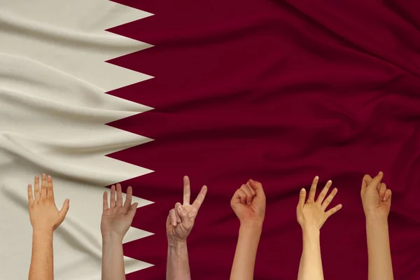 many hands raised up against the background of the national flag of Qatar on delicate shiny silk with soft draperies, the concept of the country\'s population, horizontal, close-up, copy space