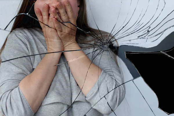 Девушка с длинными волосами закрывала лицо руками, отражение в зеркале разбитое на осколки, проблемная концепция — стоковое фото