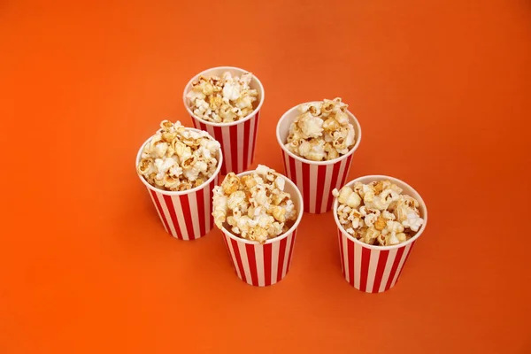 Cinco tazas de papel rojo a rayas, blanco con palomitas de maíz en una fila sobre un fondo naranja brillante, primer plano, espacio de copia, concepto de comida rápida — Foto de Stock