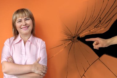 middle aged european woman smiles, arms crossed, cracks, male hand shows finger accusatory, orange background, close-up, copy space clipart
