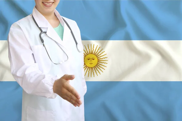 Joven hermosa niña médico con una sonrisa abierta extiende su mano para saludar en el fondo de la bandera nacional, conceptos de salud, medicina — Foto de Stock