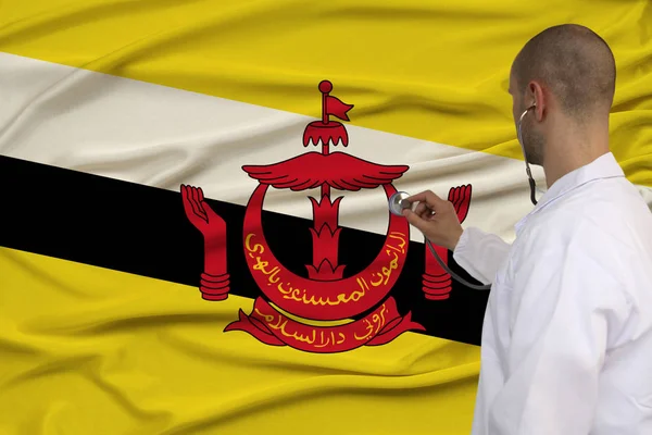 Narin parlak ipek üzerinde Brunei devletinin ulusal bayrağının arka planına karşı üniformalı bir doktorun kısmi fotoğrafı, ülkede tıbbi bakım ve sigorta kavramı, kopya aralığı — Stok fotoğraf