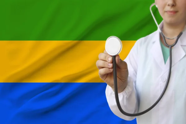 Fotografía parcial de una doctora, enfermera con un estetoscopio en uniforme en el fondo de la bandera nacional del estado de Gabón, el concepto de atención médica y seguro en el país — Foto de Stock
