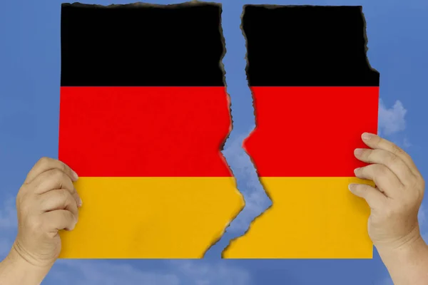 A woman holds in both hands against a blue sky torn in two cardboard with seared edges depicting the national flag of Germany, the concept of state crisis, renunciation of citizenship, destruction — Stock Photo, Image