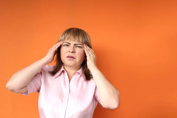Европейская женщина средних лет держит голову, оранжевый фон, концептуальная головная боль, стресс, крупный план, копировальное пространство — стоковое фото