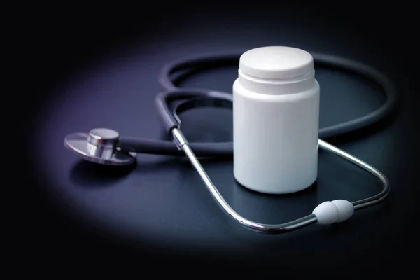 Estetoscópio médico encontra-se em um fundo preto em torno de uma lata de medicina branca vazia, forma para um designer, close-up, espaço de cópia — Fotografia de Stock